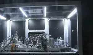 This complex machine setup is housed within a brightly lit, enclosed laboratory environment, dominated by white and grey tones.