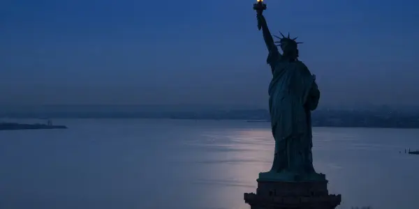 The Statue of Liberty stands illuminated at dusk, overlooking a calm harbour and distant city skyline, in shades of blue and grey.