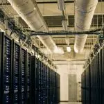 Long data centre aisle between rows of server racks with blue lights and overhead ducts