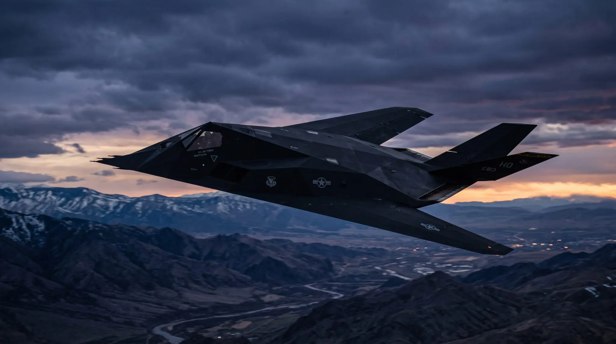 A dark F-22 Raptor fighter jet flies through a dramatic sky above rugged mountain ranges at sunset.