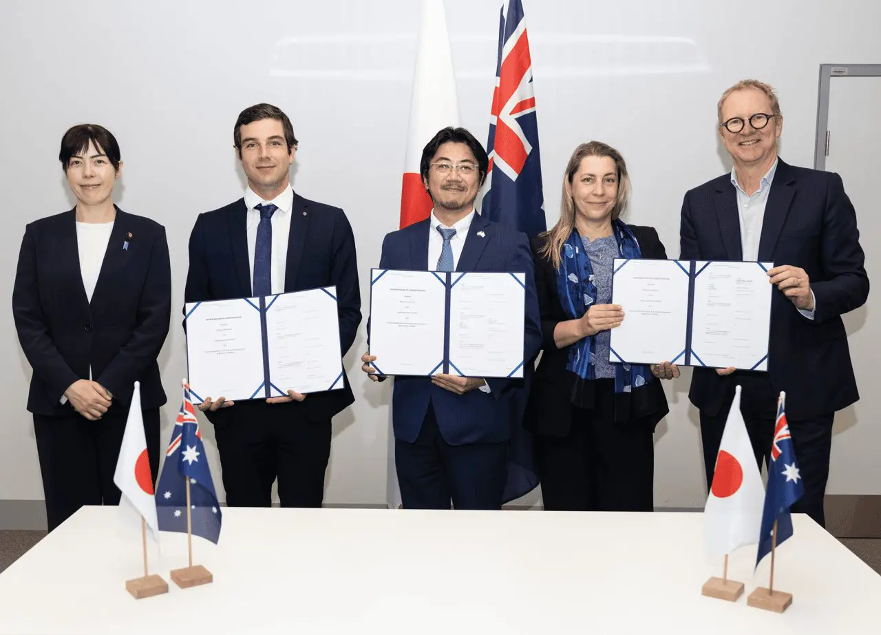 Five individuals stand around a table displaying signed documents, with flags of Japan and Australia flanking the scene.