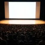 A darkened theatre is filled with seated figures facing a brightly lit, blank screen, dominated by dark wood and black