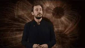 A man in a dark coat stands before a large, abstract, swirling brown and orange background. The setting is dark, with a dramatic, intense visual effect.