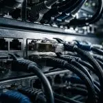 A close-up of a network switch with numerous ethernet cables connected, set in a dark server room with blue and silver tones.