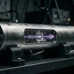 A metal atomic clock beam tube on a laboratory bench, a violet beam glowing through its window