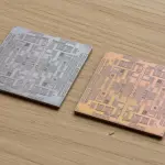 Two square circuit boards with intricate patterns rest on a light wood surface, illuminated by soft, directional lighting.