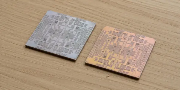 Two square circuit boards with intricate patterns rest on a light wood surface, illuminated by soft, directional lighting.