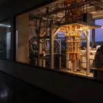 a gold dilution refrigerator seen through a lab window at dusk with a researcher at a bench