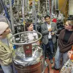 Five researchers meeting in a lab with large metallic instruments
