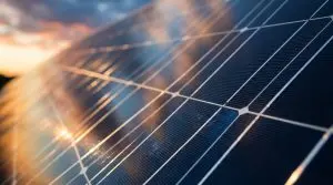 A close-up of dark blue solar panels angled towards a bright sunset, with orange and yellow light reflecting off the surface.