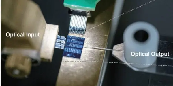 Laboratory setup showing a microchip with probe connections receiving optical input and producing optical output, illustrating integrated photonic or quantum device testing.
