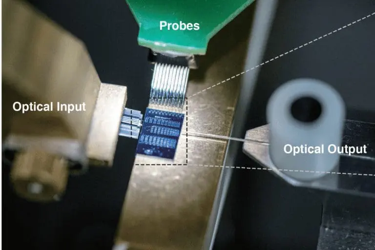 Laboratory setup showing a microchip with probe connections receiving optical input and producing optical output, illustrating integrated photonic or quantum device testing.