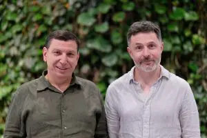 Two men stand side-by-side against a lush green wall, creating a natural and inviting backdrop.
