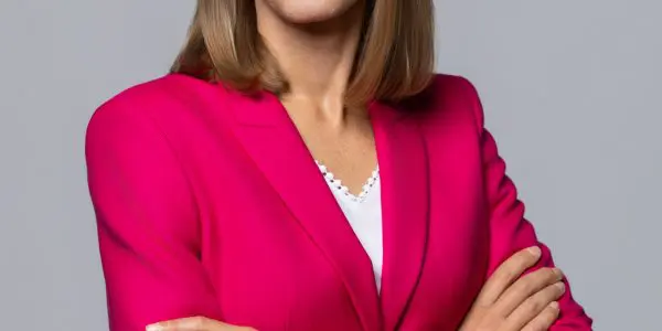 A woman in a bright pink blazer with her arms crossed stands against a grey background.