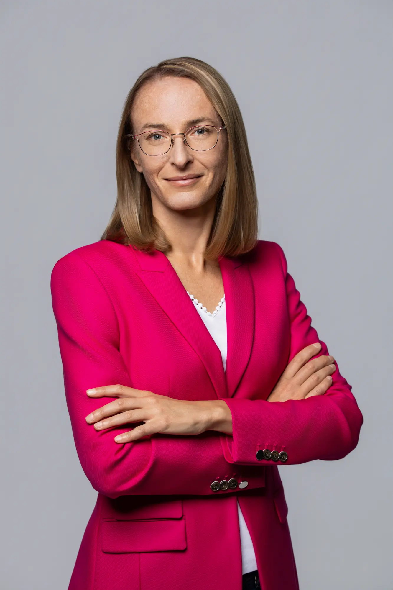 A woman in a bright pink blazer with her arms crossed stands against a grey background.