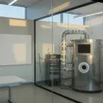 A brightly lit laboratory room with a large cylindrical device, viewed through glass, features a desk and computer.