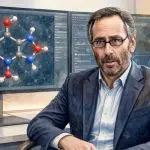 David E. Shaw seated before screens showing a molecular structure and simulation output