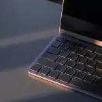 An open laptop sits on a dark surface, illuminated by warm light, with a screen displaying a dark, starry background.