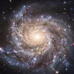 This vibrant spiral galaxy showcases intricate swirling arms against a dark, star-filled cosmic backdrop, primarily in shades of blue, pink, and gold.