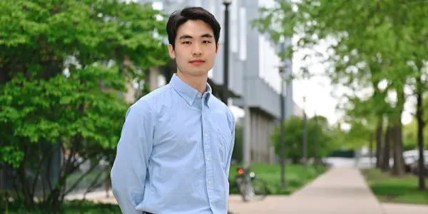 A young man stands outdoors on a sidewalk, surrounded by green foliage and a light-coloured building.