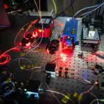 This photograph shows a complex optical experiment setup on a reflective table with numerous lasers and fiber optic cables.