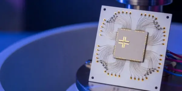 A square circuit board with gold wiring and two chips is lit with cool blue and white tones in a lab setting.
