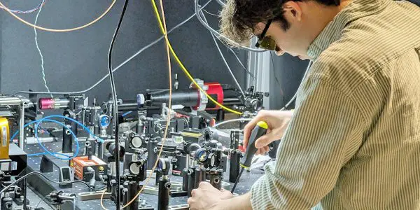 Person adjusting components on a complex optical laboratory setup with lenses, mirrors and cables arranged on a workbenc