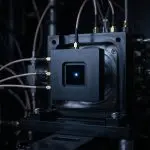 A complex, dark-toned apparatus with glowing blue light at its centre dominates the frame, surrounded by connecting tubes and wires.