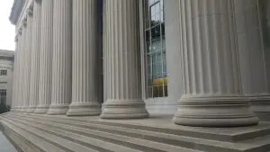 A row of tall, light-grey classical columns lines a dark exterior staircase leading to a building entrance.