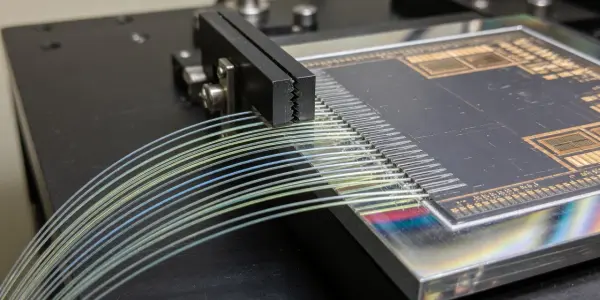 A circuit board with numerous fibre optic cables connected to it rests on a dark testing rig, with gold and silver tones.