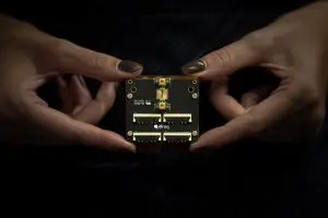 Two hands hold a small, black circuit board with various electronic components against a dark, blurred background.