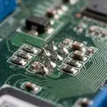 A close-up of a green circuit board with blue connectors and silver components is lit dramatically, creating sharp shadows.