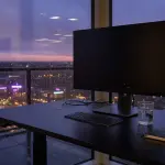 A dark desk with a monitor and notebook sits before a rainy city skyline at dusk, lit by neon.