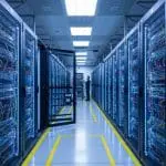 An engineer stands amid brightly lit cabled server racks in a data center corridor