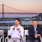 Four panelists seated on a rooftop stage at sunset, one speaking into a microphone, with the Bay Bridge and the Ferry