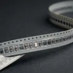 A close-up displays a strip of electronic components on a dark grey surface, with silver and white tones