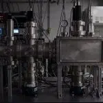 A complex scientific instrument is shown in a darkened laboratory setting with silver, black, and green equipment.