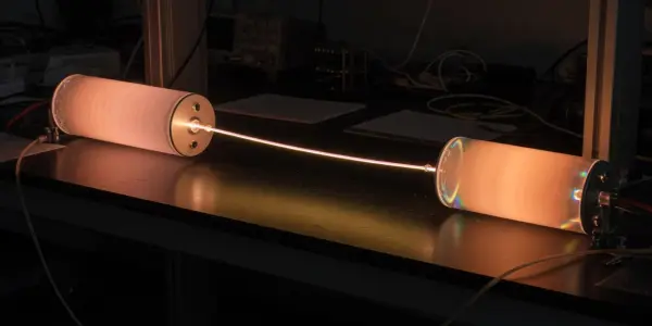 A dark laboratory bench holds a glowing optical fibre connected to cylindrical housings, illuminated with warm orange and yellow light.