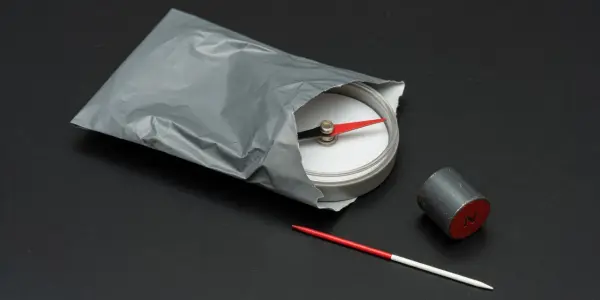 A metallic pouch, compass, magnet and needle rest on a dark grey surface, illuminated by soft, directional lighting.