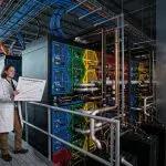 A scientist stands on a raised platform in a brightly lit data centre, reviewing a graph amidst colourful server racks.