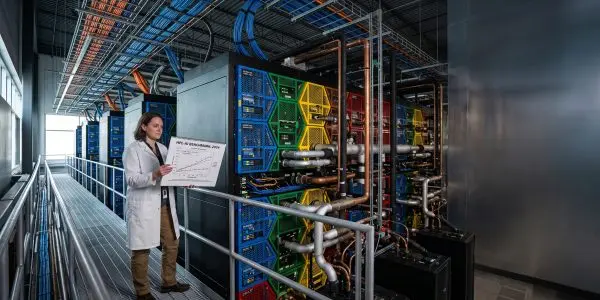 A scientist stands on a raised platform in a brightly lit data centre, reviewing a graph amidst colourful server racks.