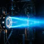 A powerful jet engine produces a bright blue exhaust stream within a darkened industrial testing chamber.