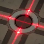 A microchip with a central circular feature illuminated by bright red laser beams against a dark background.