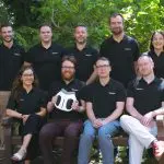A group of people in dark clothing pose outdoors amongst greenery on a sunny day.