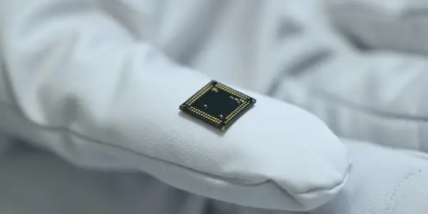 A small, dark-coloured microchip rests on a gloved hand, set against a dark background, with soft, diffused lighting.
