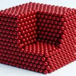 A cube constructed from many small, shiny red spheres rests on a white surface, bathed in soft light