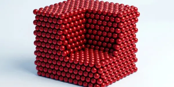 A cube constructed from many small, shiny red spheres rests on a white surface, bathed in soft light
