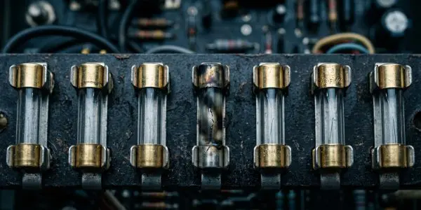 A close-up of vintage glass fuses in a dark, metallic electrical panel dominates the frame, using blues and golds