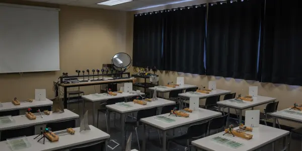 A darkened lab space contains rows of workstations, each with equipment on a green mat, lit by overhead lights.