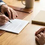 Two pairs of hands at a desk, one signing a document with a fountain pen